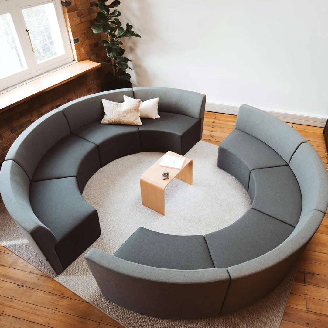 The Konfurb Arco Series by Home Office Space NZ presents an exceptional design with its contemporary, circular seating arrangement featuring dark gray upholstered sections arranged on a round, light-colored rug. A light wood coffee table, adorned with a book and a small plant, takes center stage. The space is accented by wooden floors and brick walls with windows adorning the side.