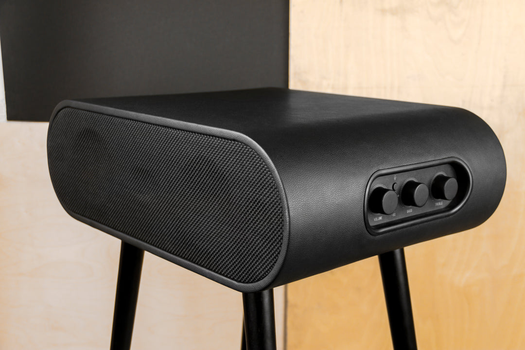 A sleek black speaker with rounded edges from Home Office Space NZ, the MySound Jazz Speaker Side-Table features three front control knobs and wireless charging capabilities. It elegantly stands against a light wood panel background with a dark rectangular section highlighting its modern design.