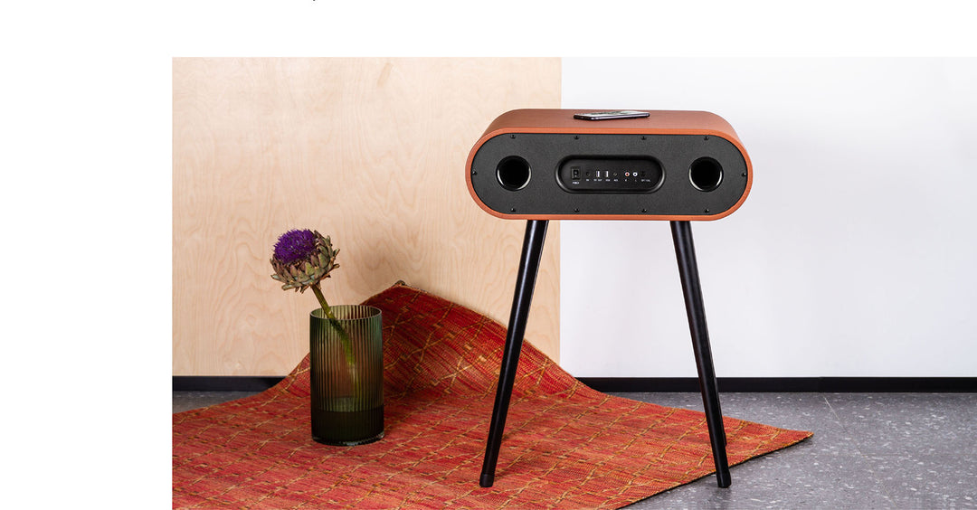 The MySound Jazz Speaker Side-Table by Home Office Space NZ is a retro-style floor speaker with a wooden finish and black legs, featuring dual side speakers, a central control panel, and wireless charging for smartphones. It’s displayed next to a green vase with purple flowers on an orange patterned rug.