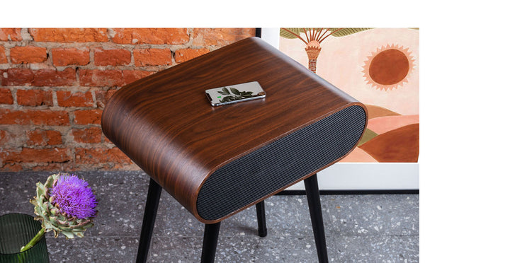 A MySound Jazz Speaker Side-Table by Home Office Space NZ, with an oval wooden design and black legs, stands on a tile floor. A leaf-patterned item tops it. The backdrop shows a brick wall and framed sun/landscape art, while a purple flower adds charm to the scene.